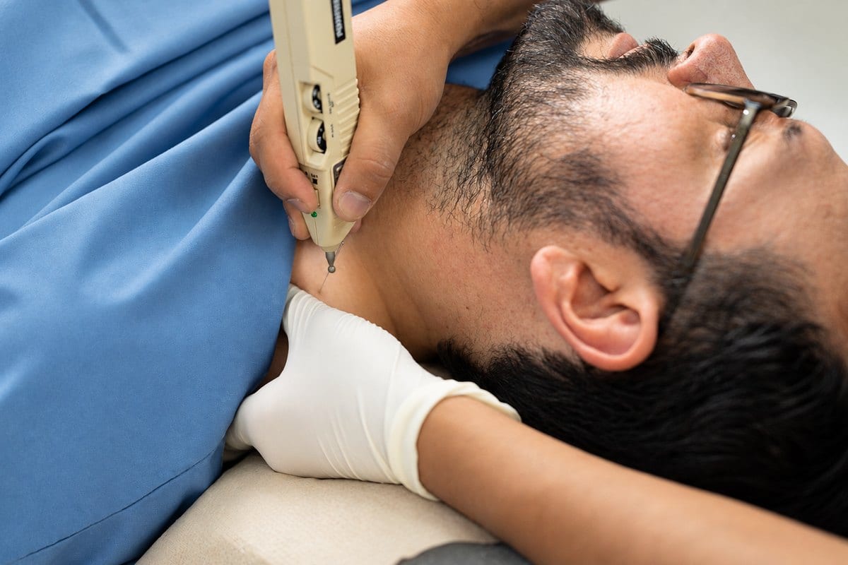 Primer plano: Juan Mesías apoya un dispositivo de estimulación sobre el cuello de un paciente recostado, sosteniendo la zona con la mano enguantada.