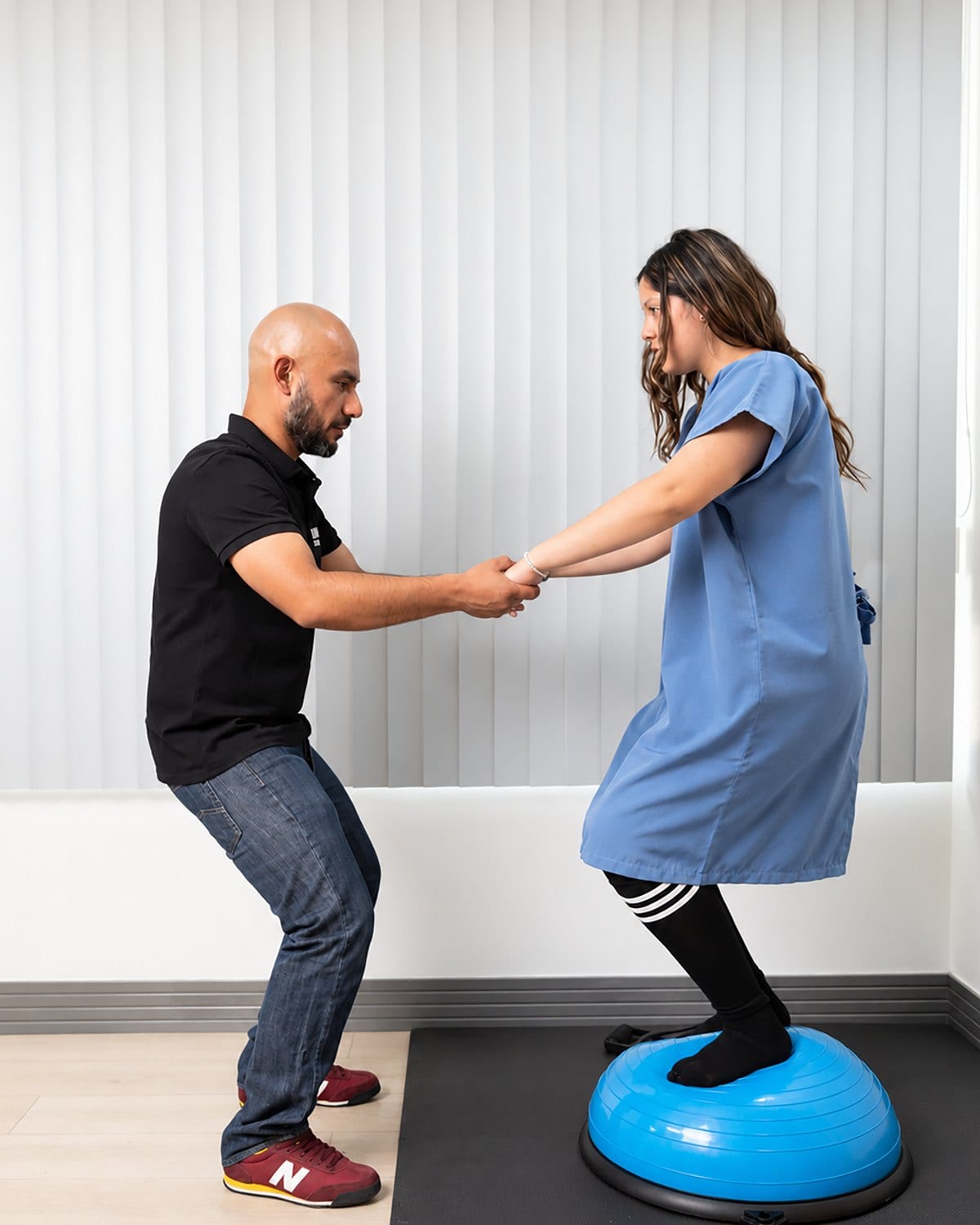 Juan Mesías sostiene las manos de una paciente que hace un ejercicio de equilibrio de pie sobre un bosu, en el consultorio.