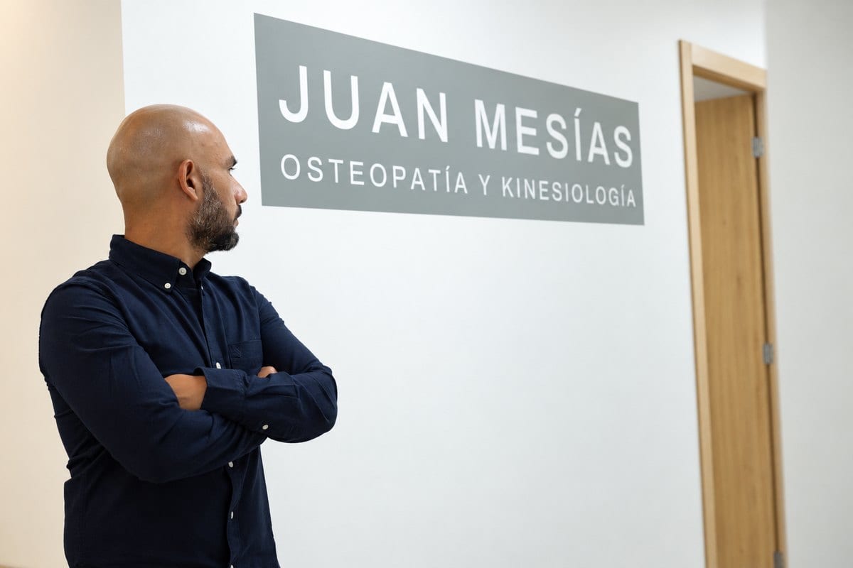 El rótulo del consultorio —«Juan Mesías · Osteopatía y Kinesiología»— en la pared del pasillo, con Juan de pie al lado.